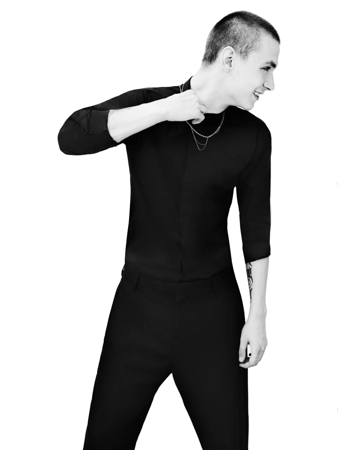 Black and white shot of a cropped-hair man in a black shirt and trousers, laughing and tugging his collar, for Dazed and Confused.