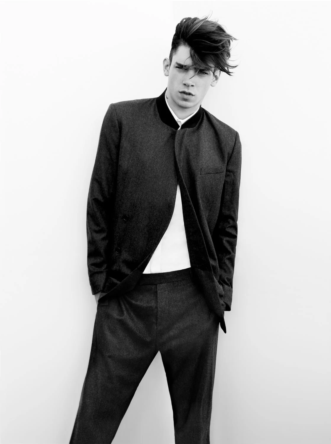 Black and white shot of a male model with tousled hair in a dark collarless suit and white shirt, for Dazed and Confused.
