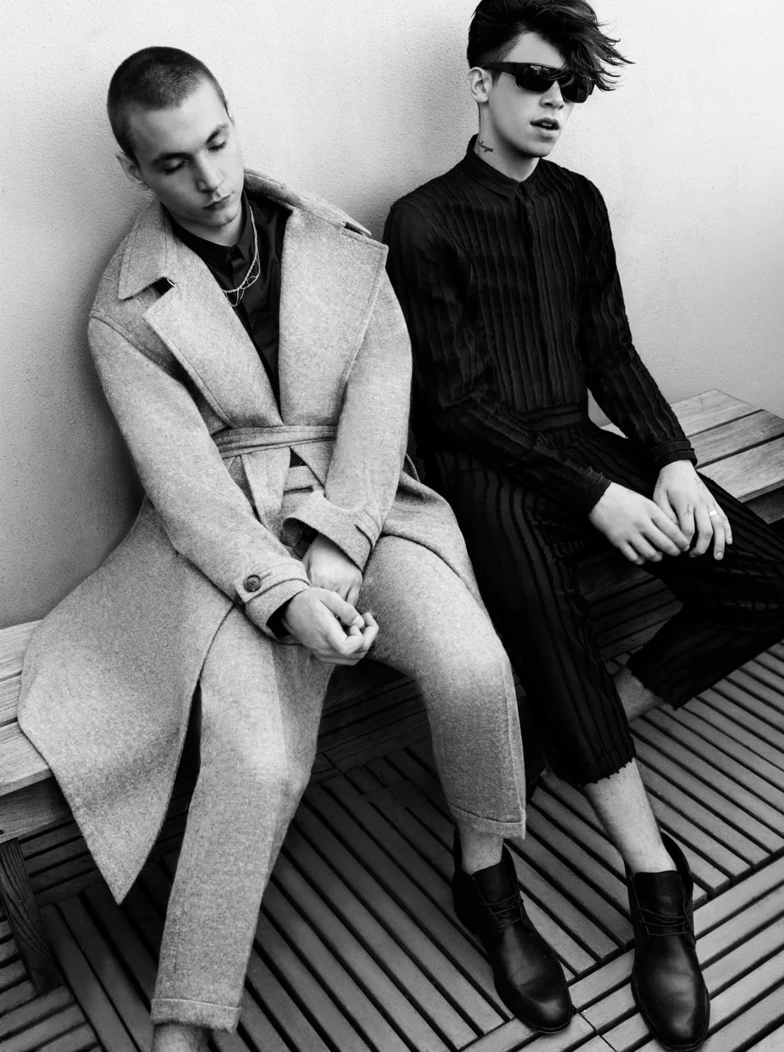 Black and white shot of two young men on a slatted wooden bench, one in a belted wool coat, one in sunglasses, for Dazed and Confused.