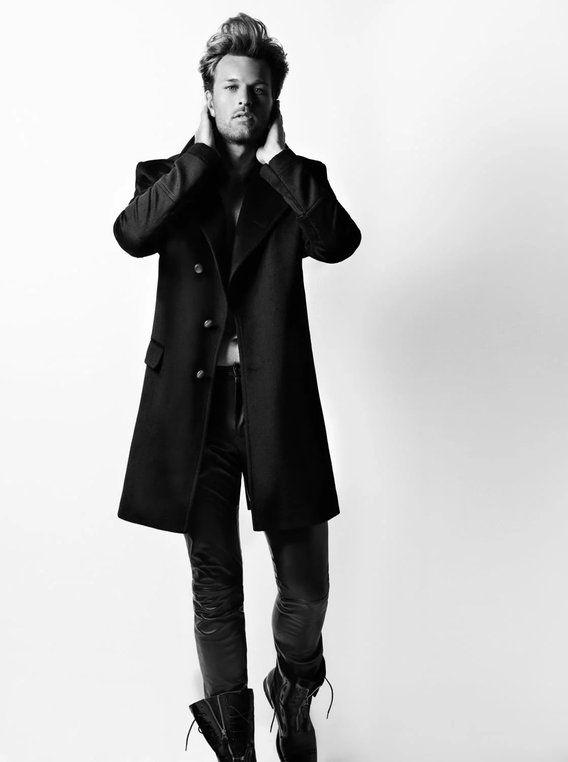 Black and white shot of a man in a black double-breasted overcoat, leather trousers and boots, hands at his neck, for Out Magazine.