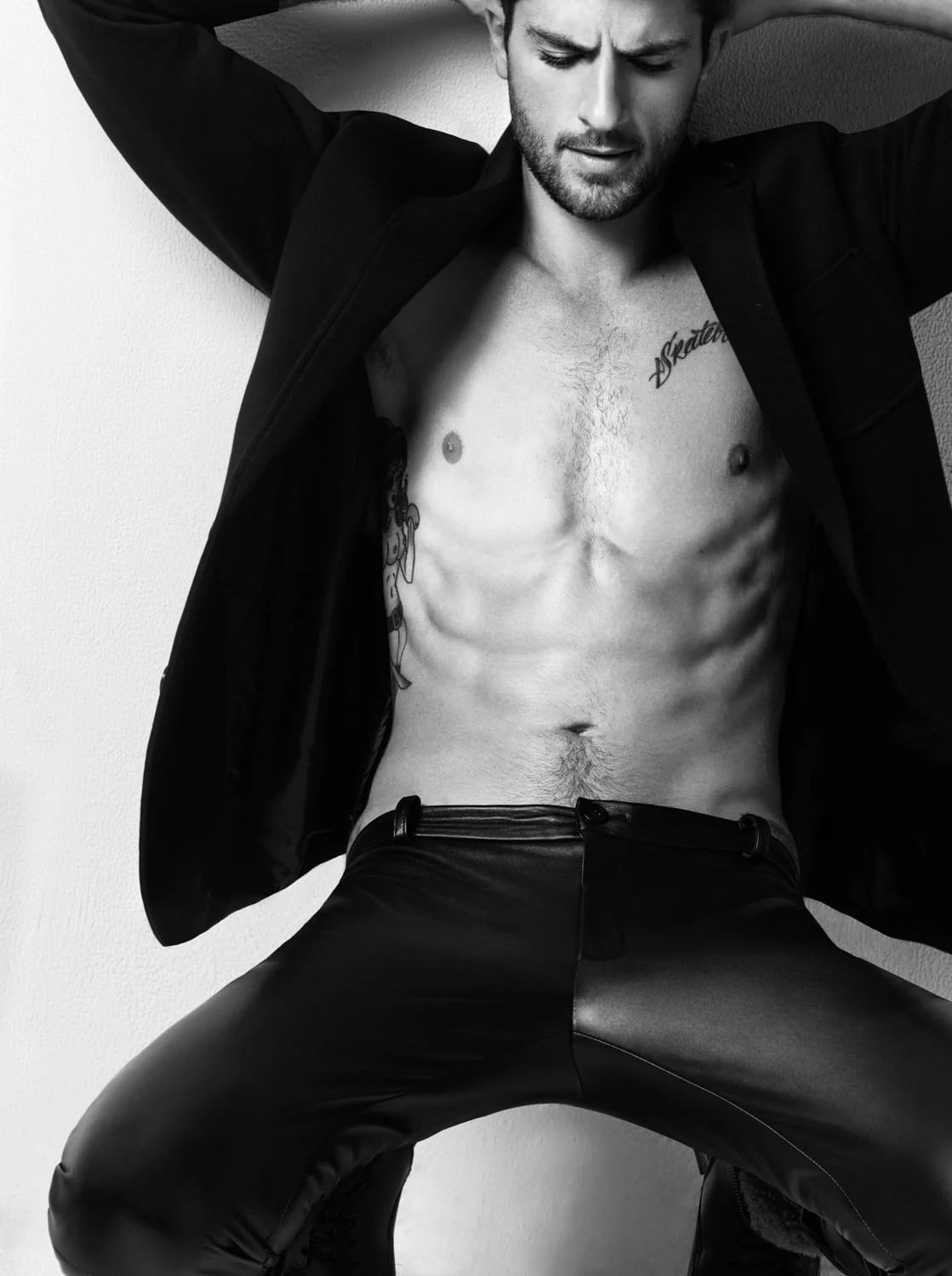 Black and white shot of a shirtless tattooed man in an open black blazer and leather trousers against a wall, for Out Magazine.
