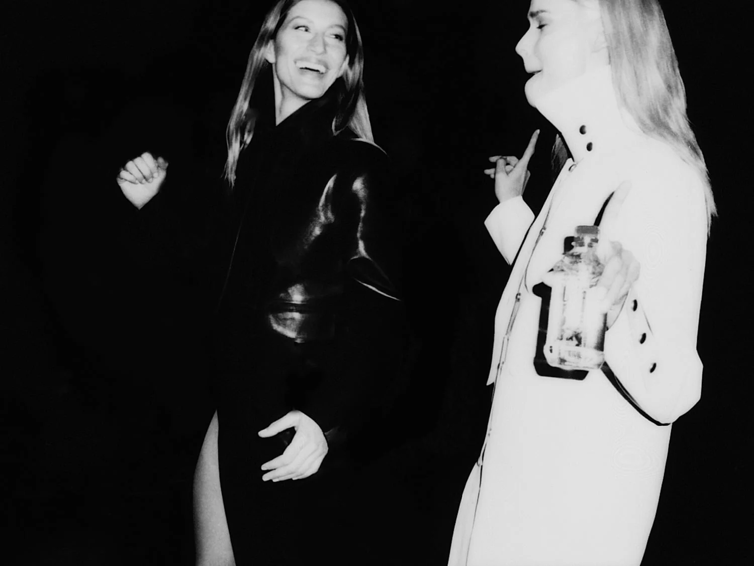 Black-and-white flash photo of two laughing women, one in a black leather coat, one in a white coat holding a bottle.