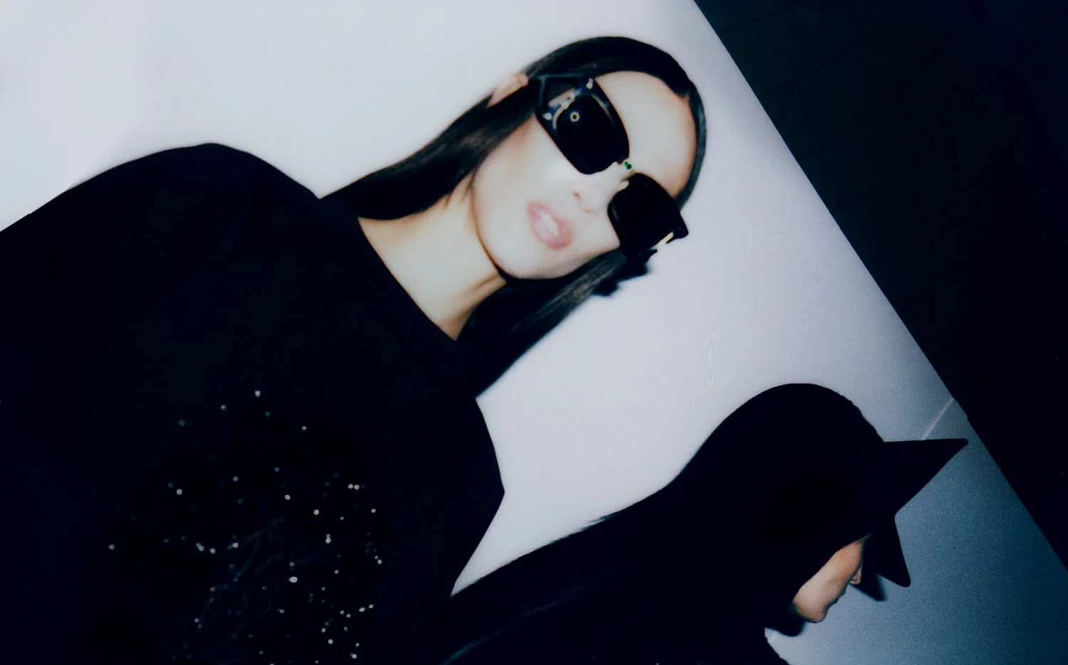 Tilted flash shot of a model in oversized black sunglasses and a sparkling black top against a white wall, for The New York Times Style Magazine.