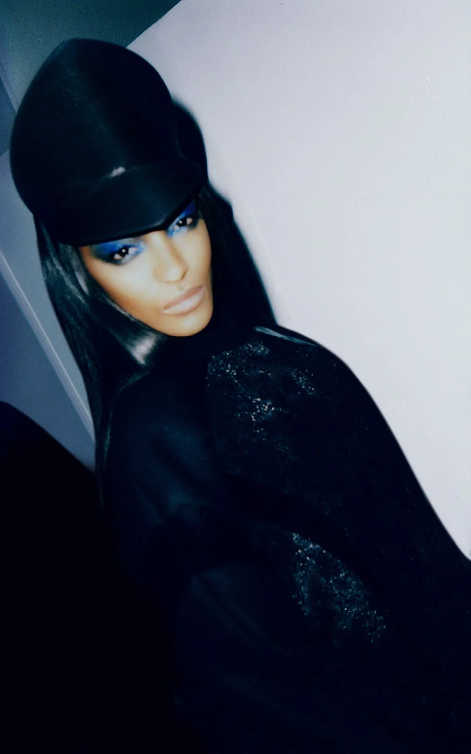 Model in a black peaked cap, blue eyeshadow and sparkling black coat leaning on a white wall, for The New York Times Style Magazine.