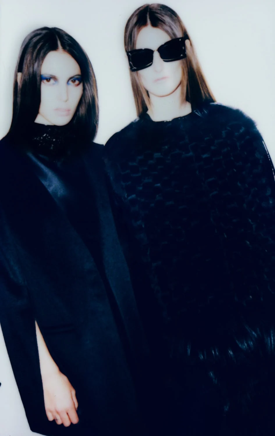 Two models in black satin and woven fur coats, one in dark sunglasses, against a white wall, for The New York Times Style Magazine.