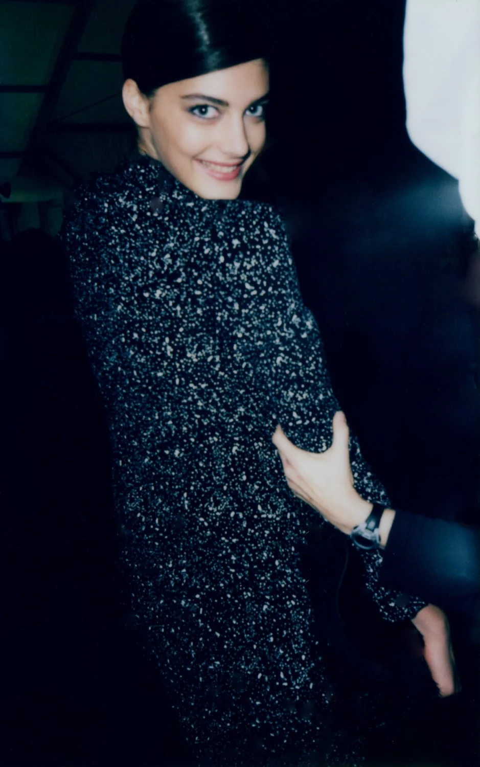 Smiling model with slicked-back hair in a black speckled knit dress, a hand adjusting her sleeve backstage, for The New York Times Style Magazine.