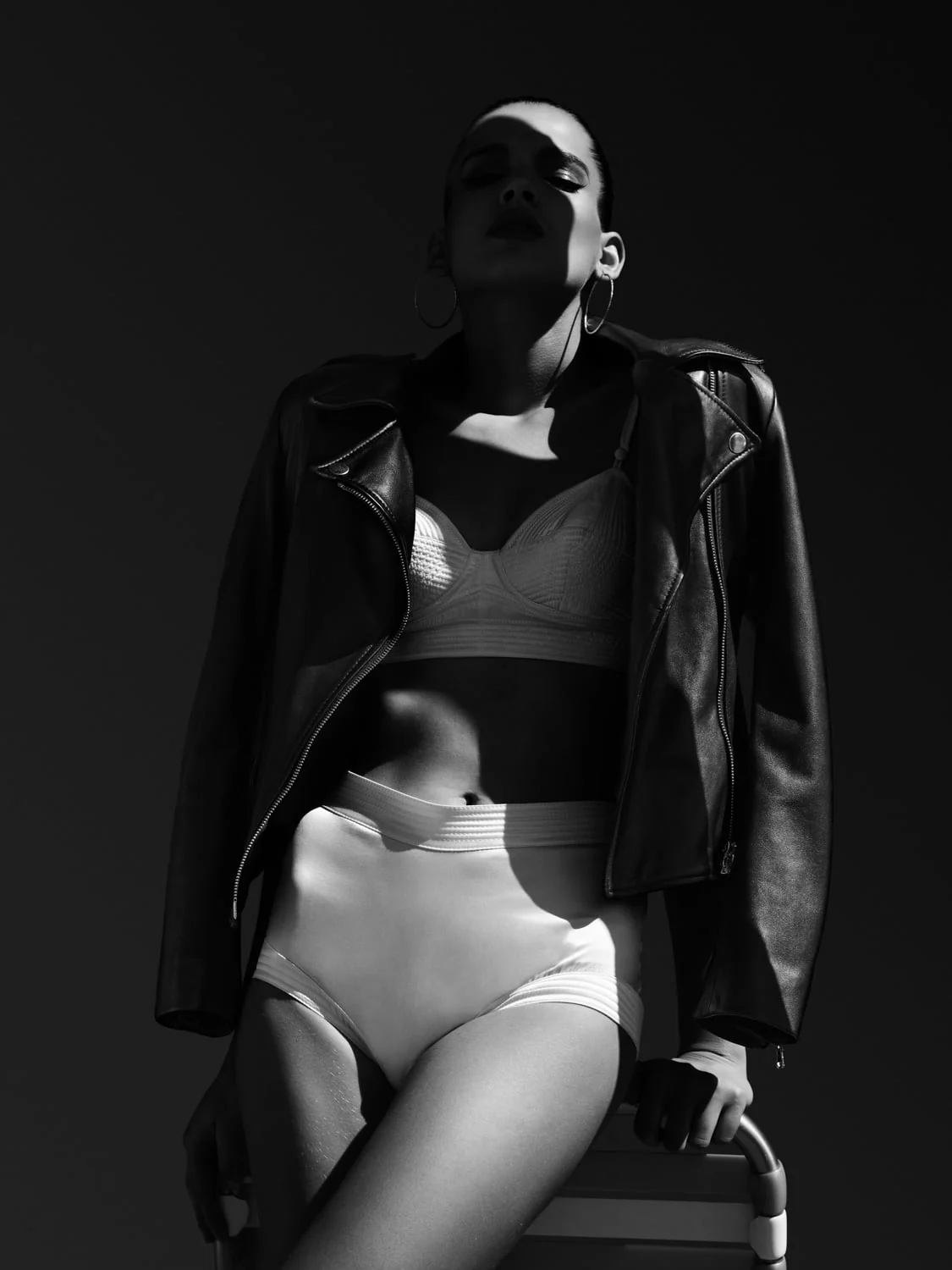 Black and white shot of a model in a leather biker jacket, white bra and briefs, hoop earrings, in hard light, for Amica. Photographed by Cameron Krone.