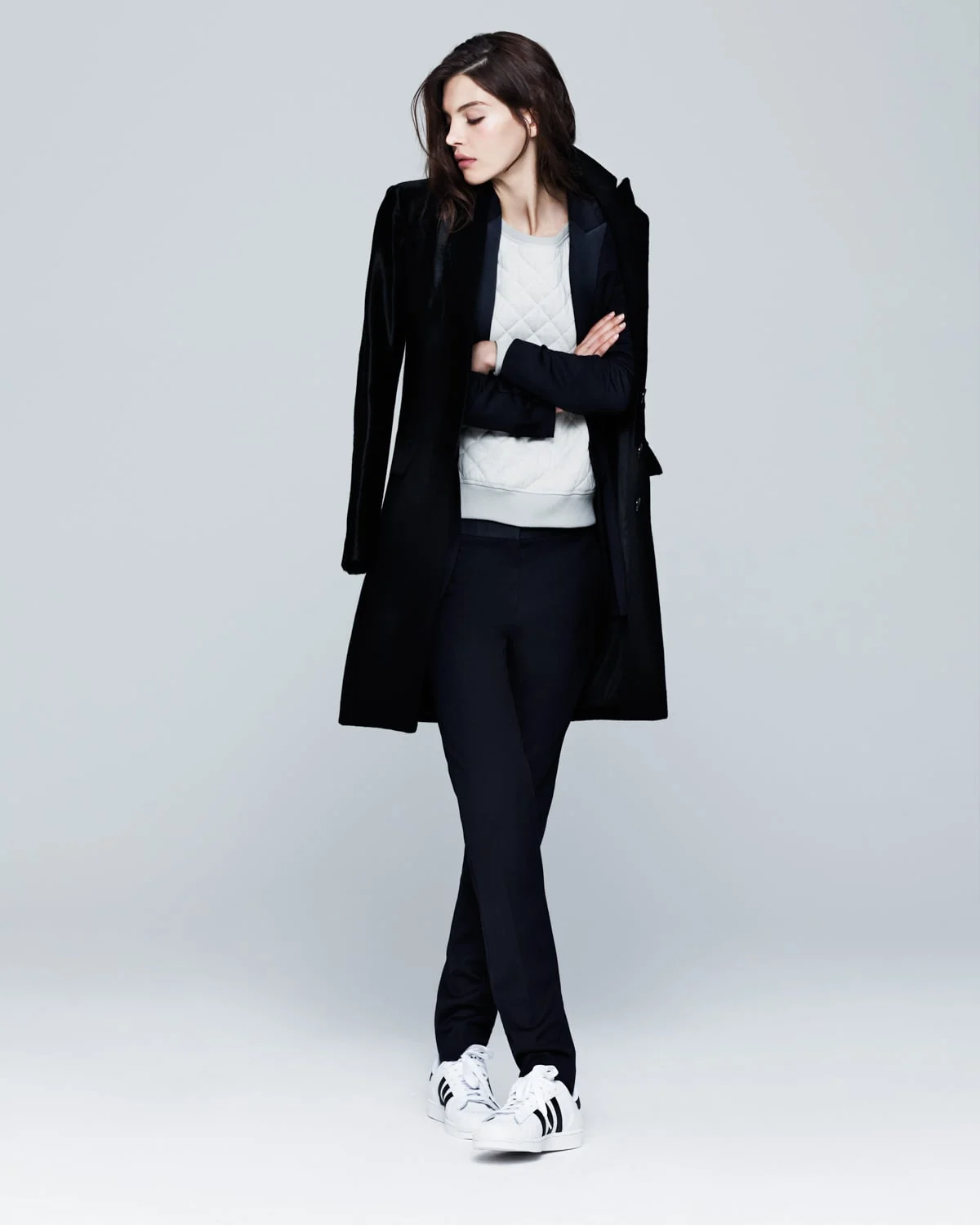Model in a long black velvet coat, white quilted sweatshirt, black trousers and white sneakers against a grey backdrop.