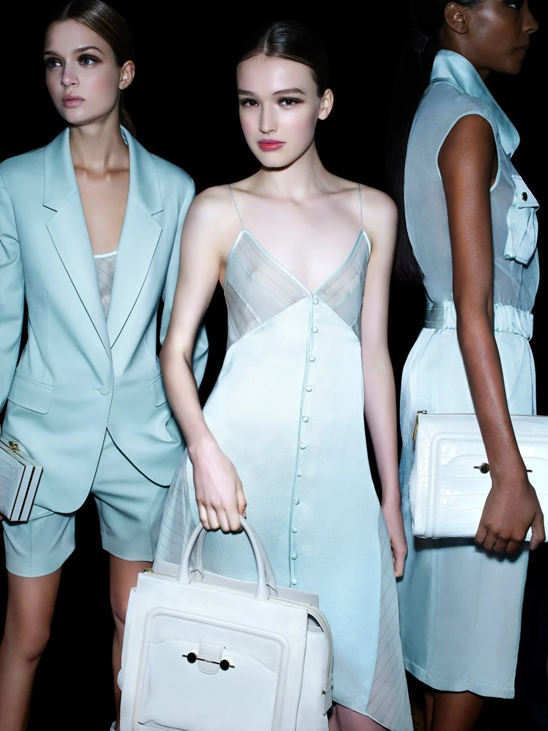 Three models in pale mint suit, slip dress and shirtdress holding white handbags against black, for Jason Wu.