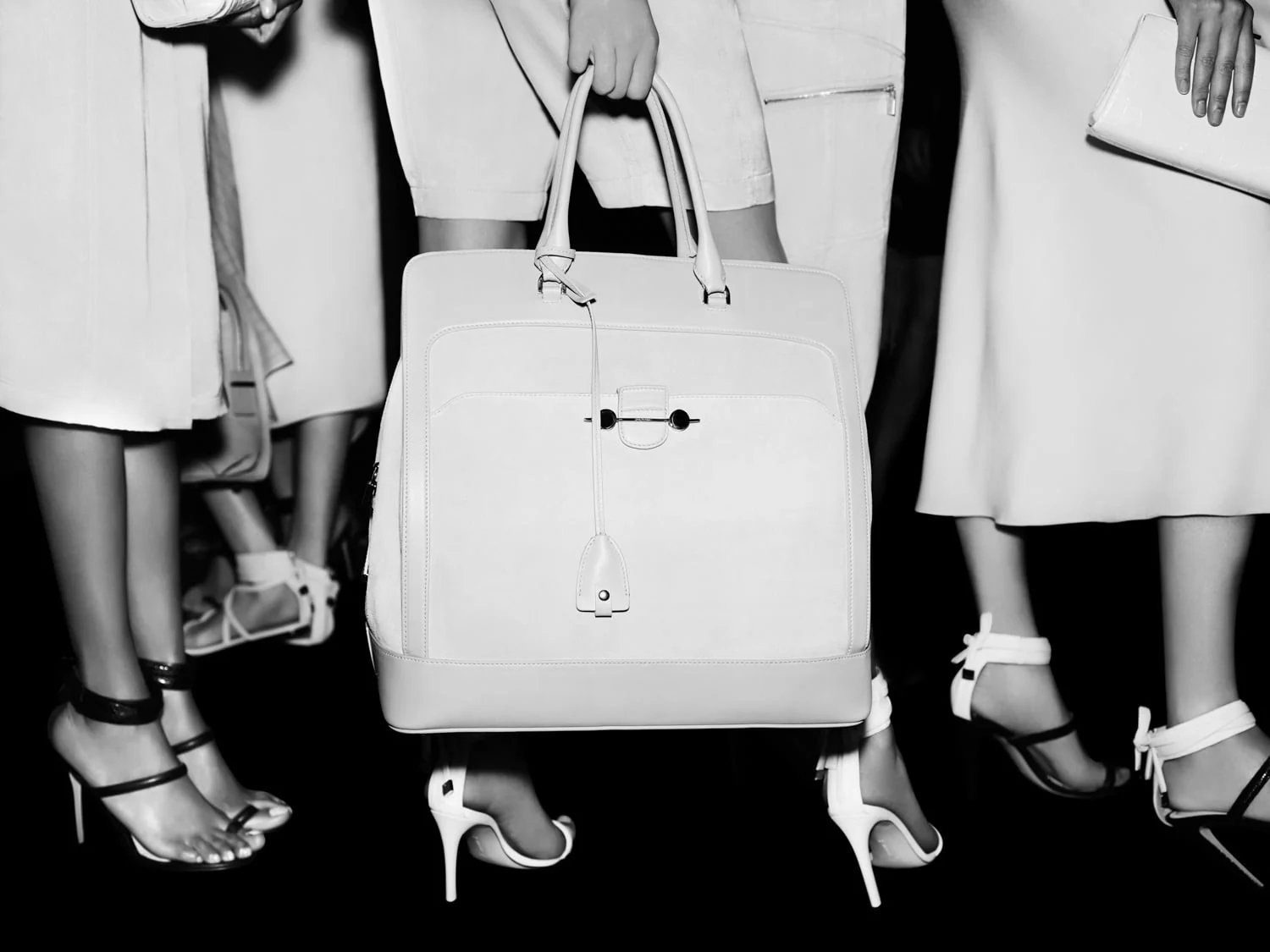 Black-and-white shot of a model holding a white leather tote among legs in strappy heels and pale dresses, for Jason Wu.
