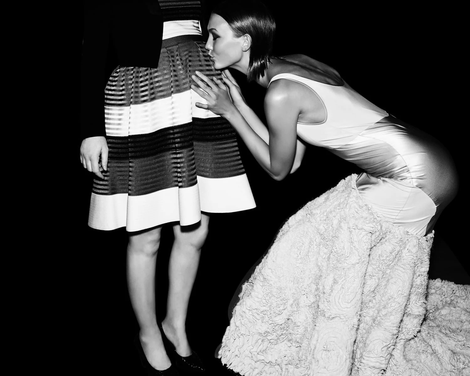 Black and white shot of a model in a satin gown with ruffled train kissing another model's striped skirt, for Jason Wu.