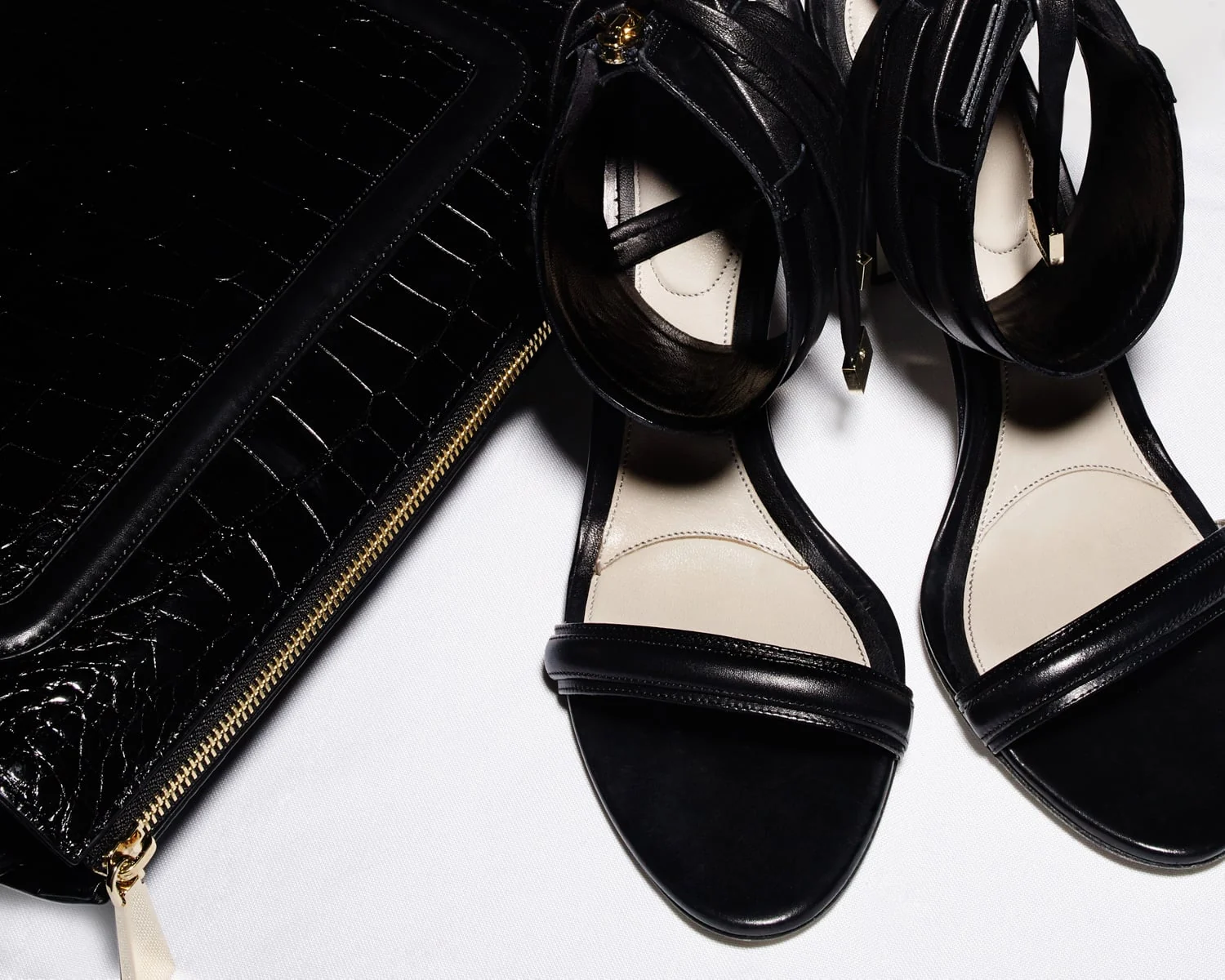 Black leather ankle-cuff sandals beside a glossy black croc-embossed clutch with gold zipper on white, for Jason Wu.