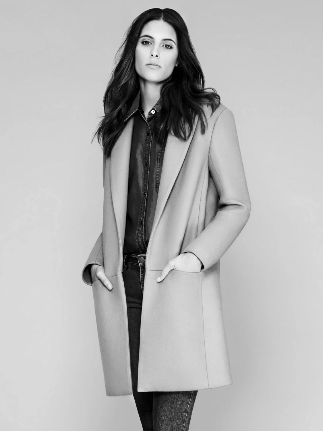 Black-and-white studio portrait of a woman in a long wool coat, denim shirt and jeans, hands in her pockets.