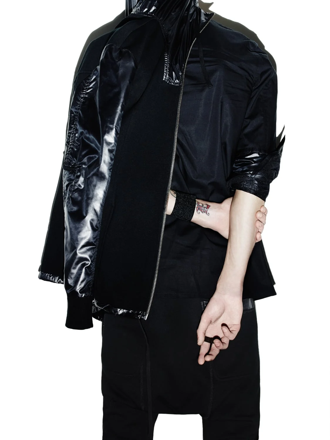 Model in layered black nylon and knit zip jacket with arms crossed behind, tattooed wrist visible, for Helmut Lang.