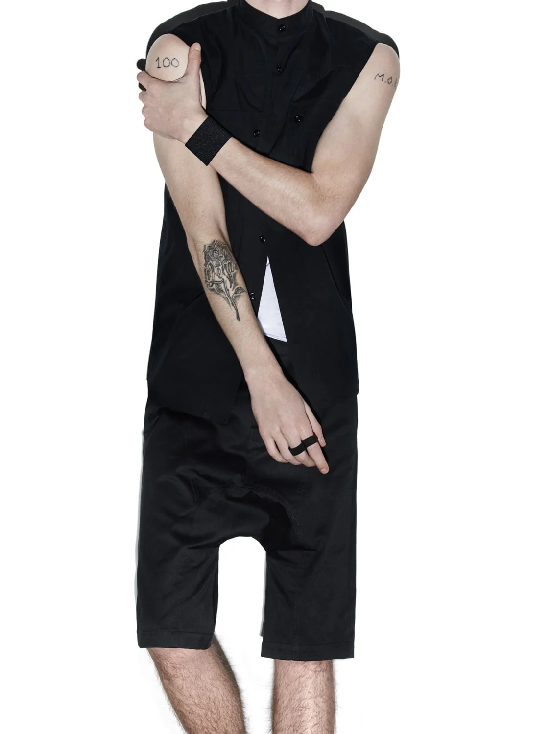 Model in a black sleeveless band-collar shirt and drop-crotch shorts, arms crossed, tattooed forearm, for Helmut Lang.