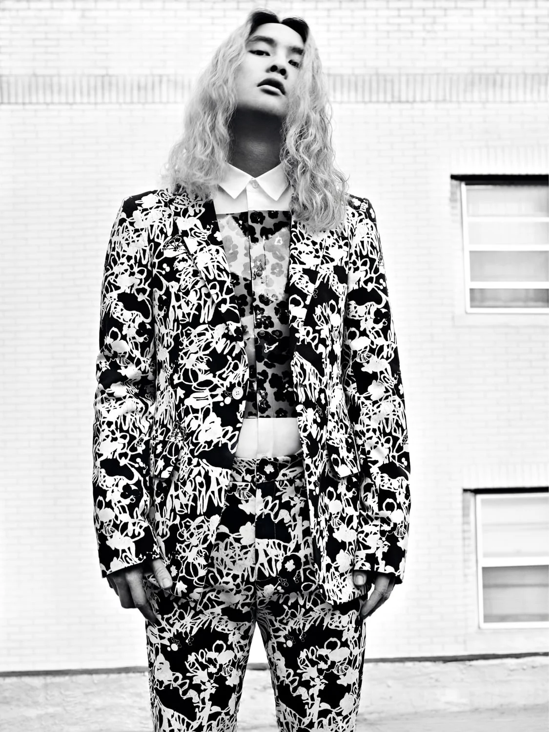 Black and white shot of a model with long wavy blond hair in a graffiti print suit against a white brick wall.