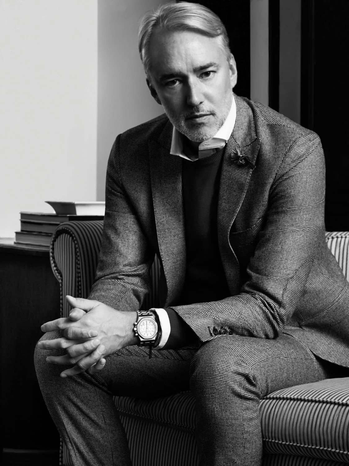 Black and white portrait of a grey-haired man in a tweed suit and wristwatch, seated on a striped armchair, hands clasped.