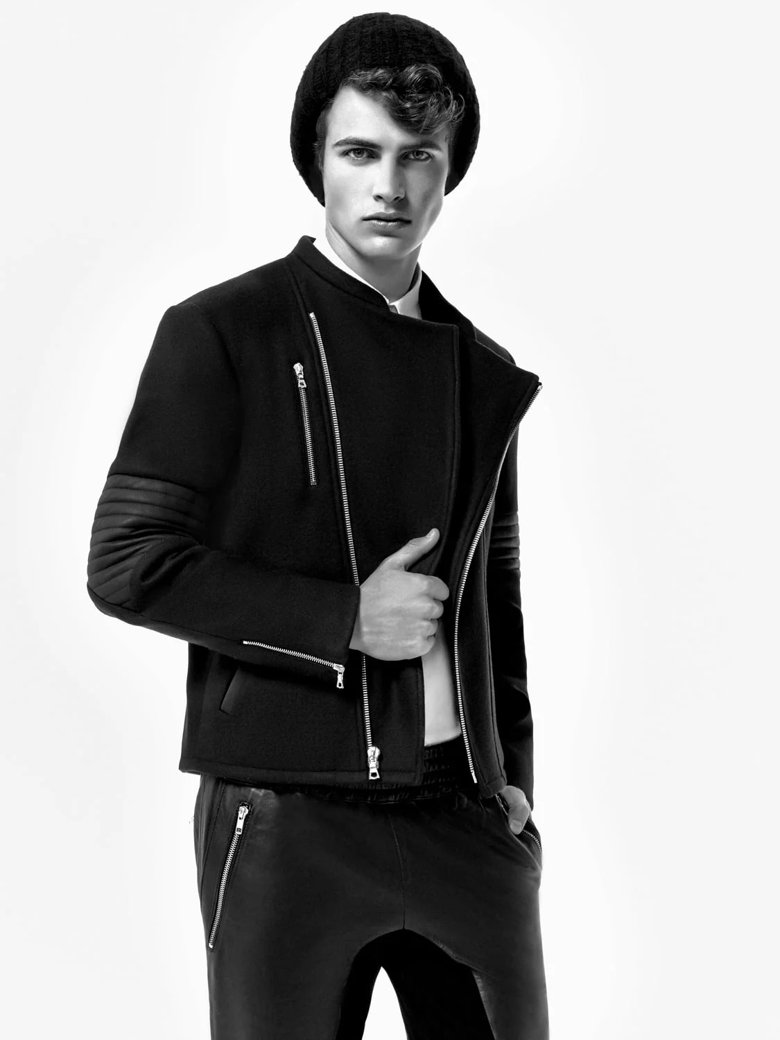 Black and white shot of a male model in a beanie, zippered wool moto jacket and leather trousers, for Saks Fifth Avenue.
