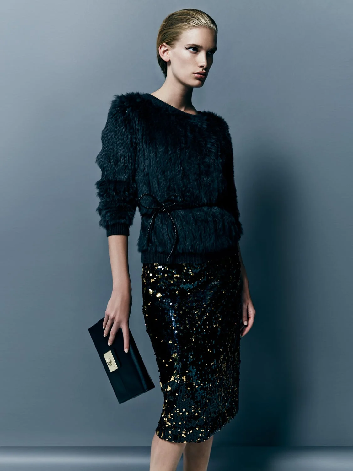 Model in a black fur sweater and gold-flecked sequin pencil skirt holding a black clutch on grey, for Saks Fifth Avenue. Photographed by Cameron Krone.