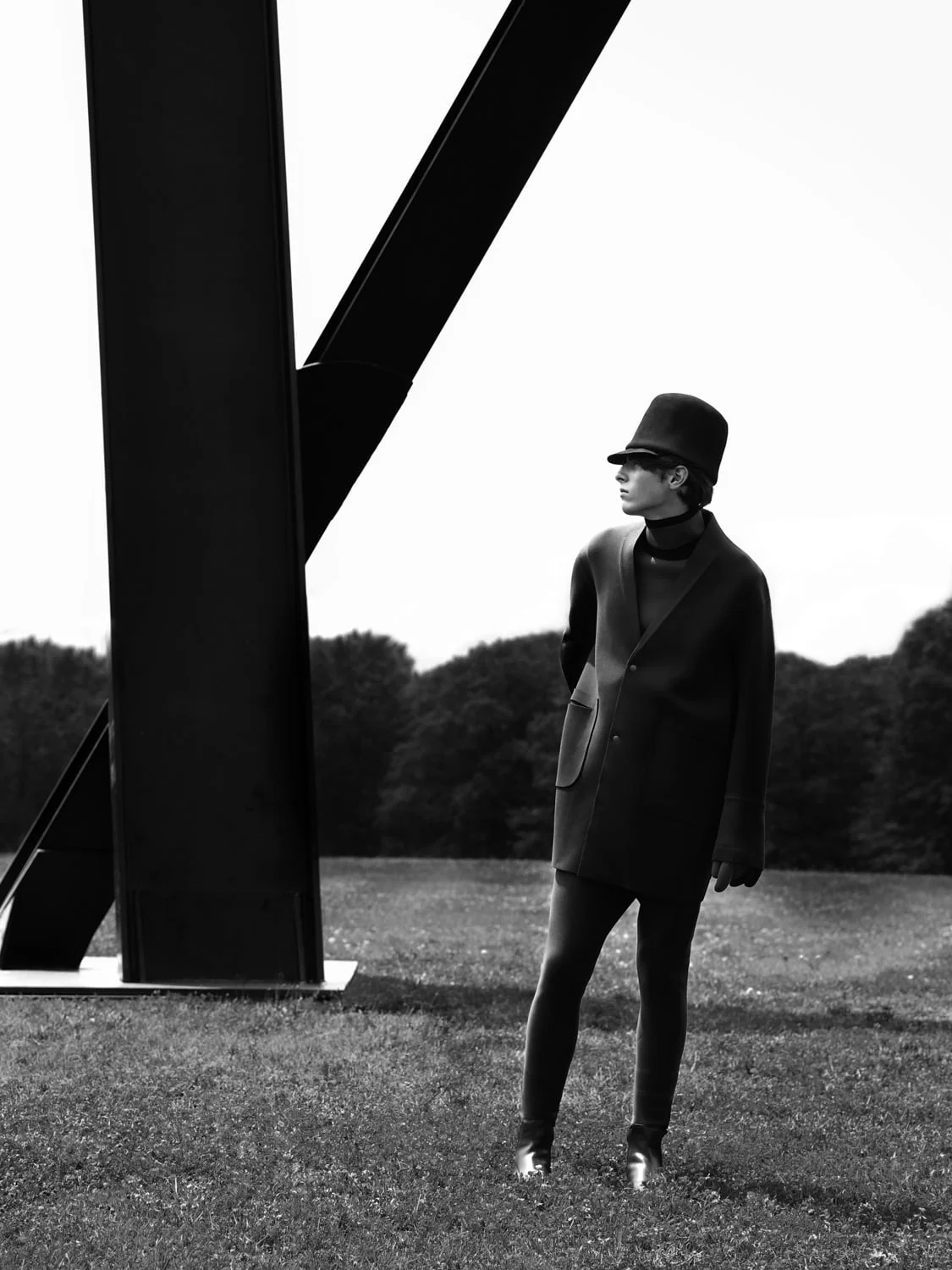Black and white shot of a model in a felt hat and oversized coat standing on grass beside a large steel sculpture.