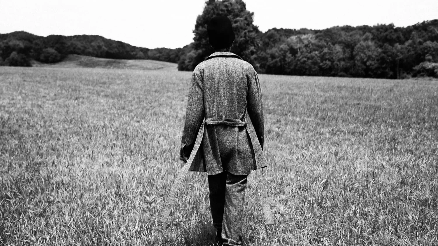 Black and white shot of a person in a belted tweed coat walking away across a grassy field toward trees, for Hercules. Photographed by Cameron Krone.
