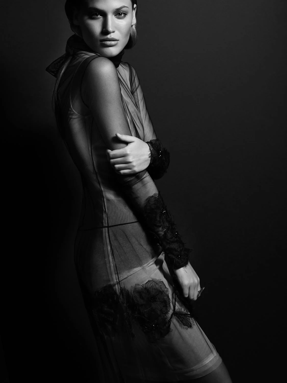 Black and white portrait of a model in a sheer high-neck gown with black floral lace appliqué, posed against a dark backdrop.