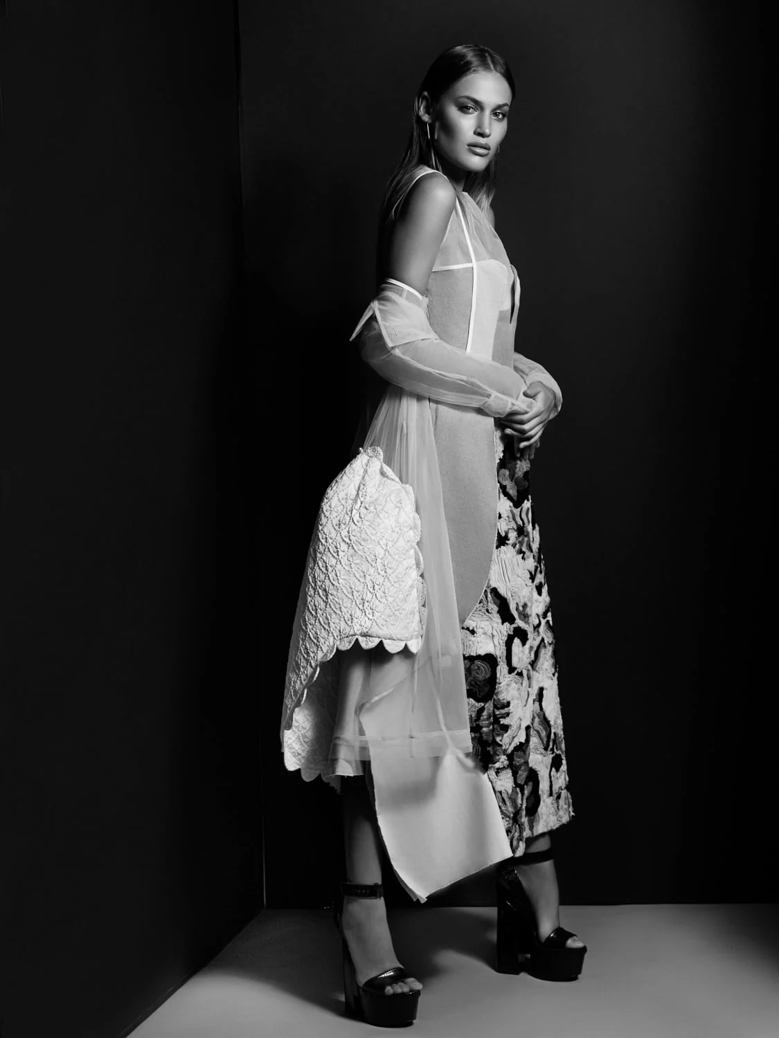Black-and-white portrait of a model in a sheer layered dress, quilted jacket and platform sandals against a dark backdrop.