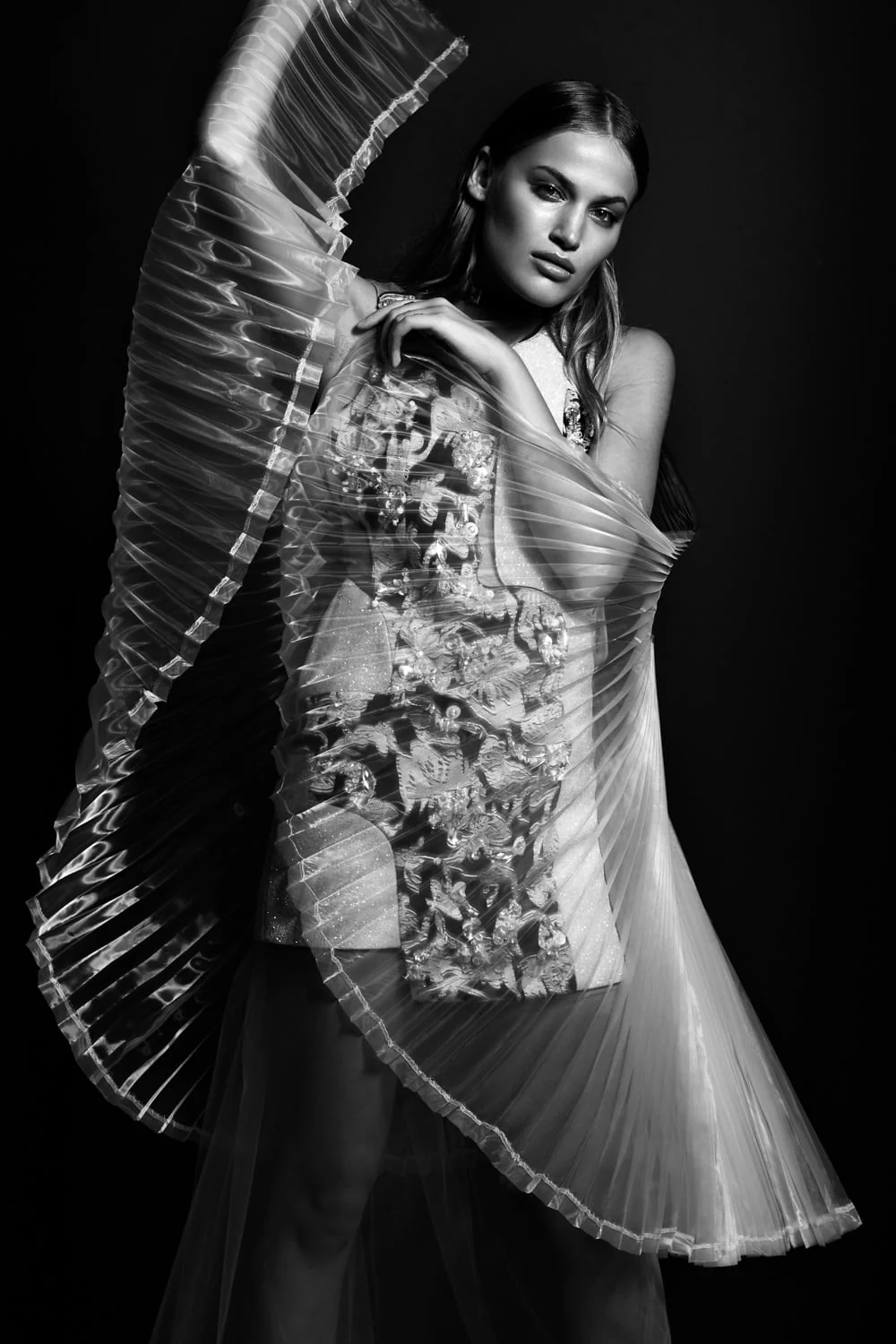 Black-and-white portrait of a model holding a sheer pleated organza cape over an embroidered dress against a dark backdrop.