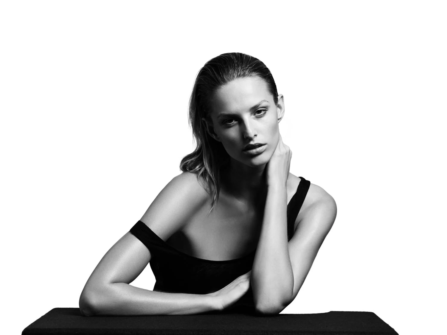 Black and white portrait of a woman with slicked-back hair in an off-shoulder black top, leaning on a black table.