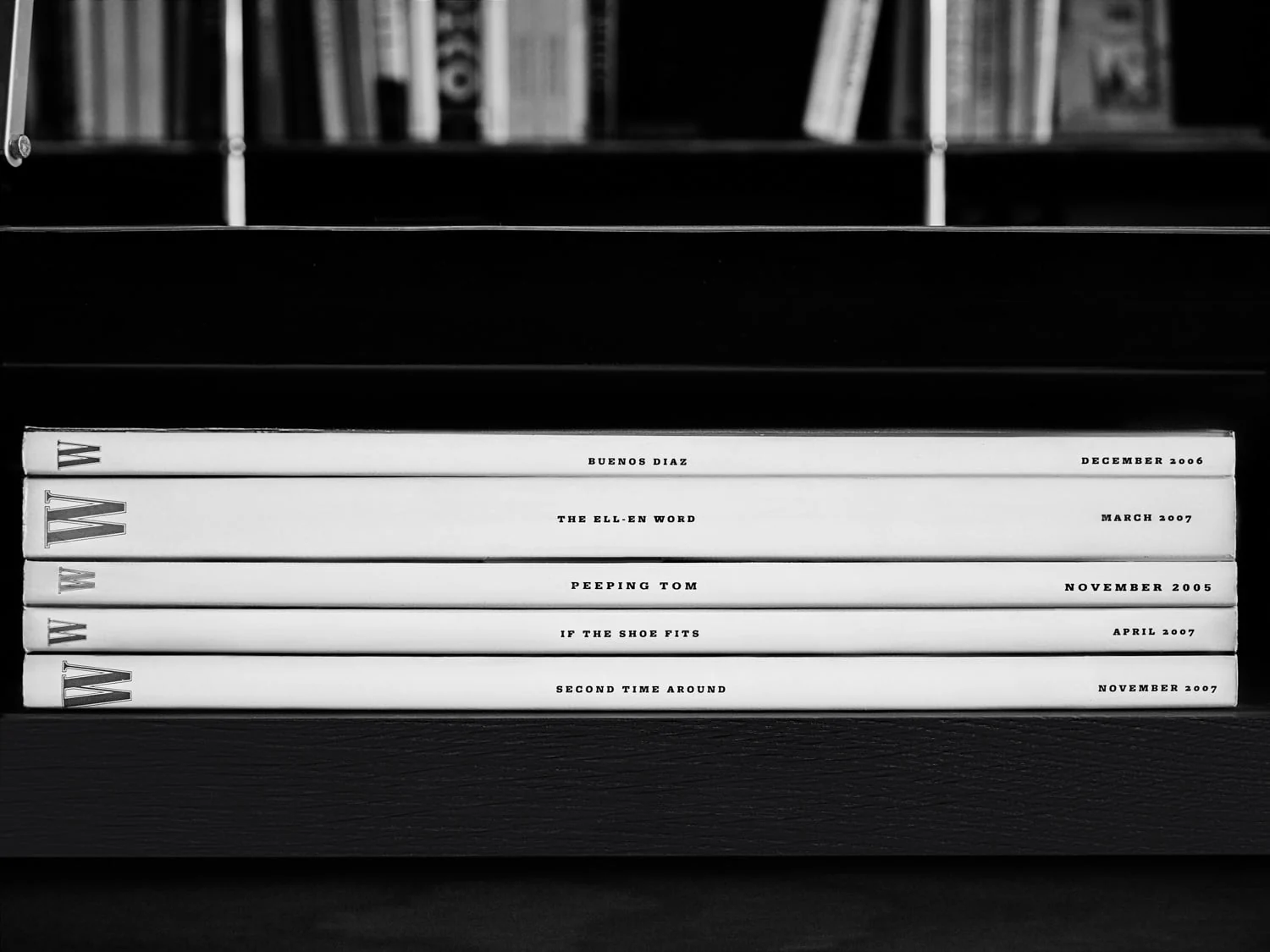 Black and white stack of five W magazine issues from 2005 to 2007, spines facing out on a dark bookshelf.