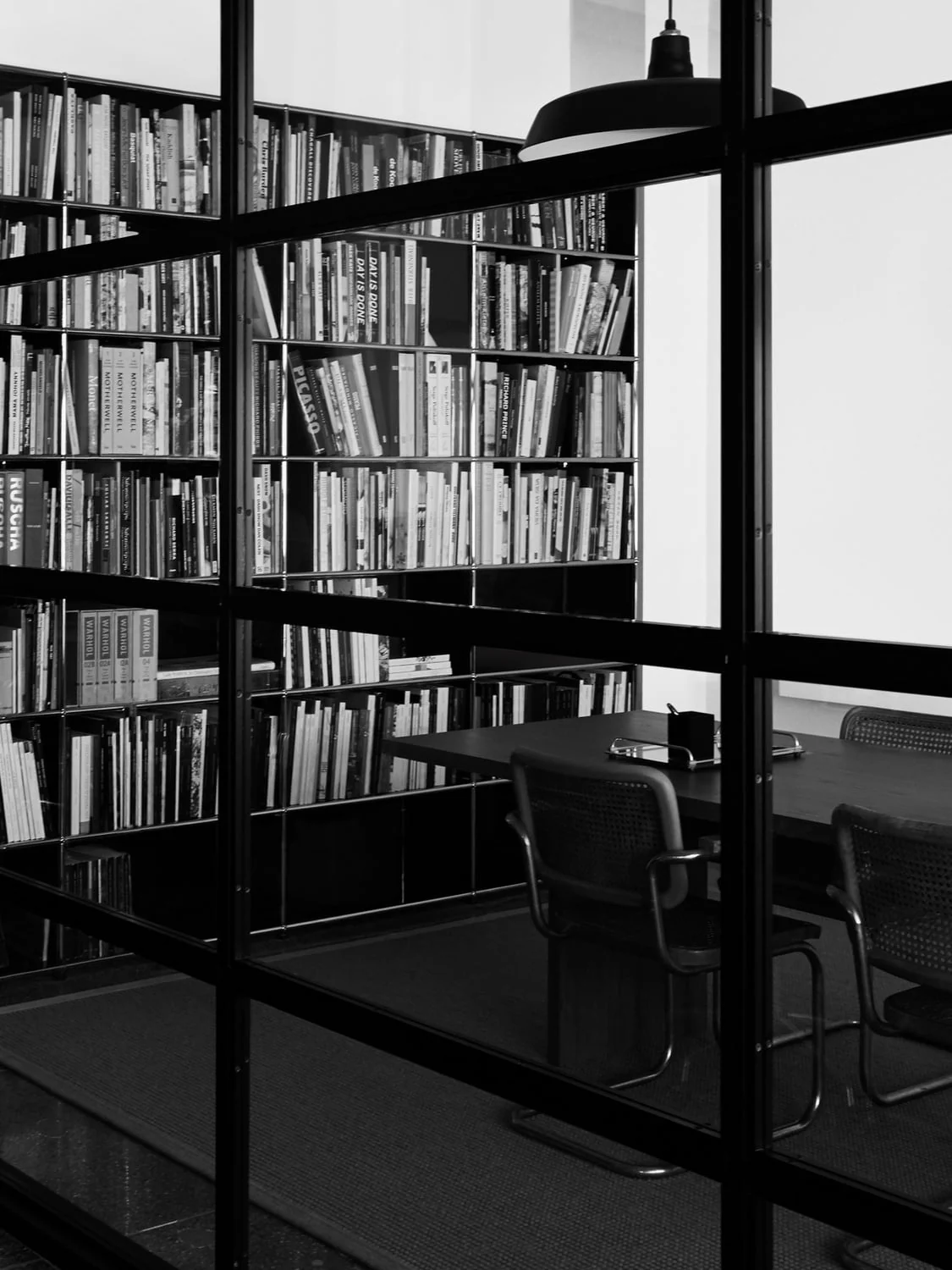 Black and white view through a steel-framed glass wall of a meeting room with tall bookshelves, cane chairs and a pendant lamp.