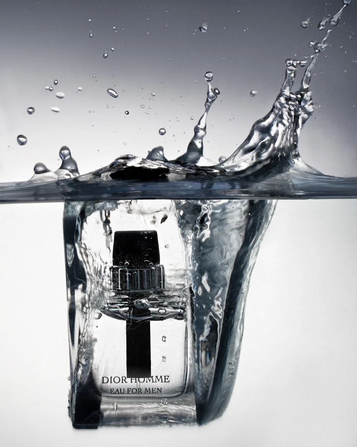 Dior Homme Eau for Men bottle plunging into water with a splash and droplets against a grey backdrop, for Dior.