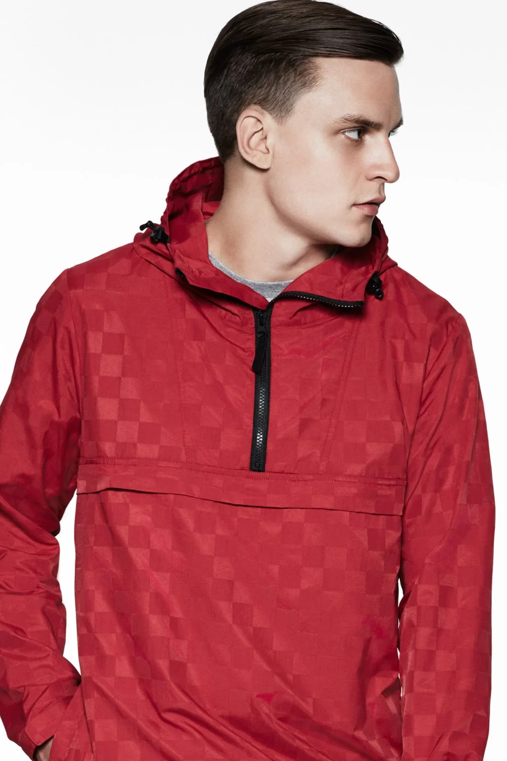 Male model in a red tonal checkerboard half-zip anorak with hood, looking aside against white, for Saturdays NYC.