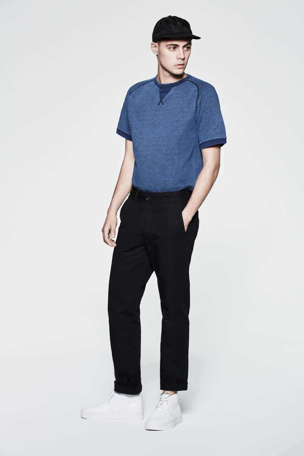 Model in a blue short-sleeve raglan sweatshirt, black chinos, black cap and white sneakers on grey, for Saturdays NYC.