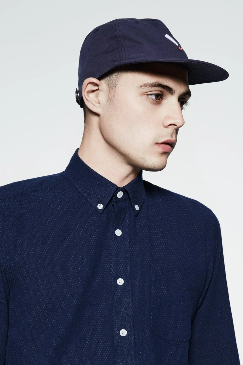 Male model in a navy six-panel cap and navy pin-dot button-down shirt against a pale grey backdrop, for Saturdays NYC. Photographed by Cameron Krone.