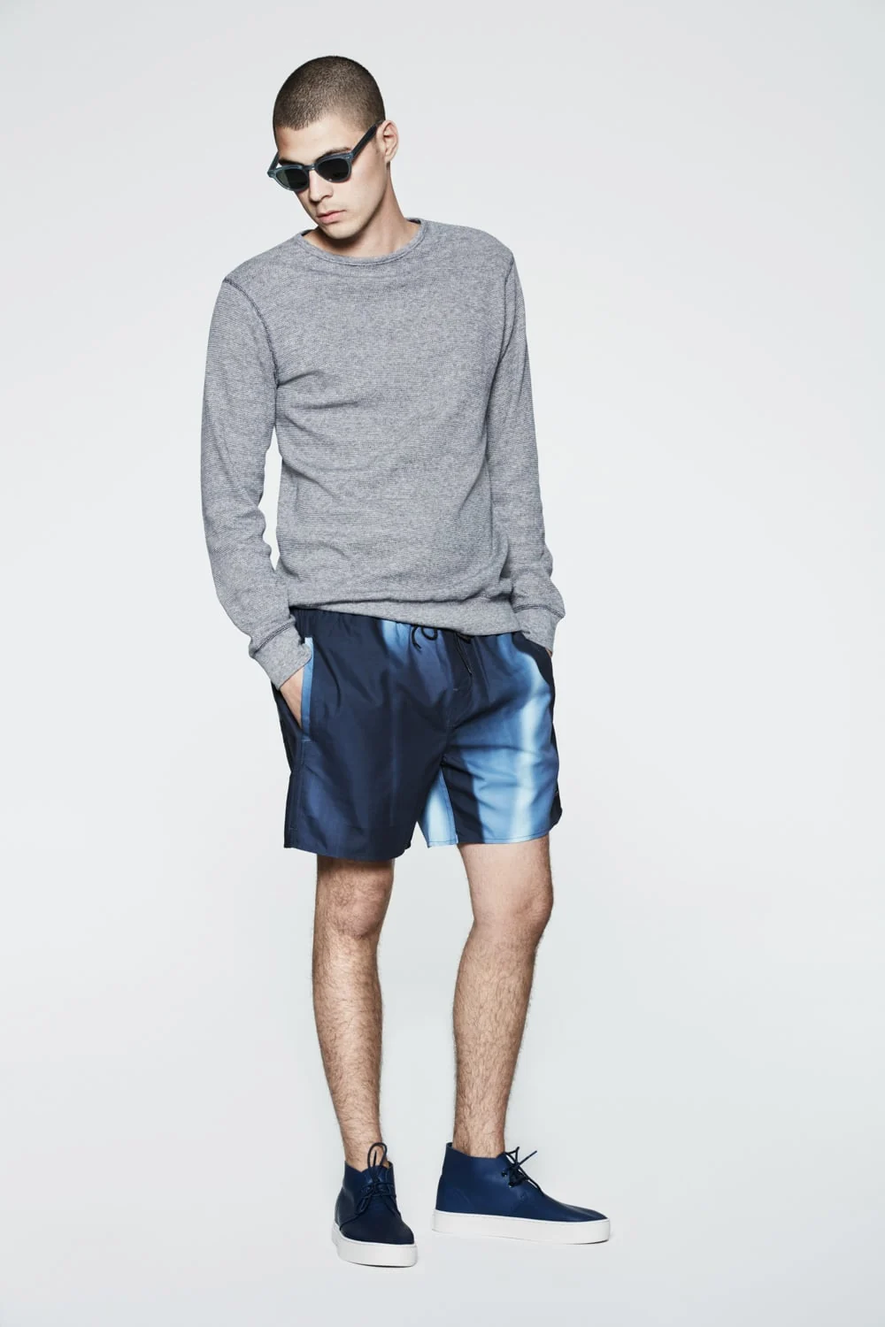 Male model in a grey knit sweater, blue printed swim shorts, sunglasses and navy sneakers on white, for Saturdays NYC.