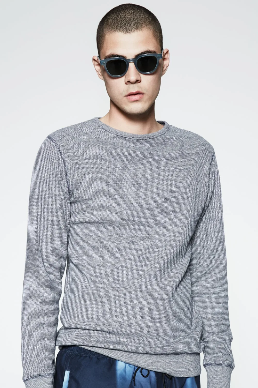 Model with buzz cut in a grey knit crewneck sweater, dark sunglasses and printed shorts against white, for Saturdays NYC.