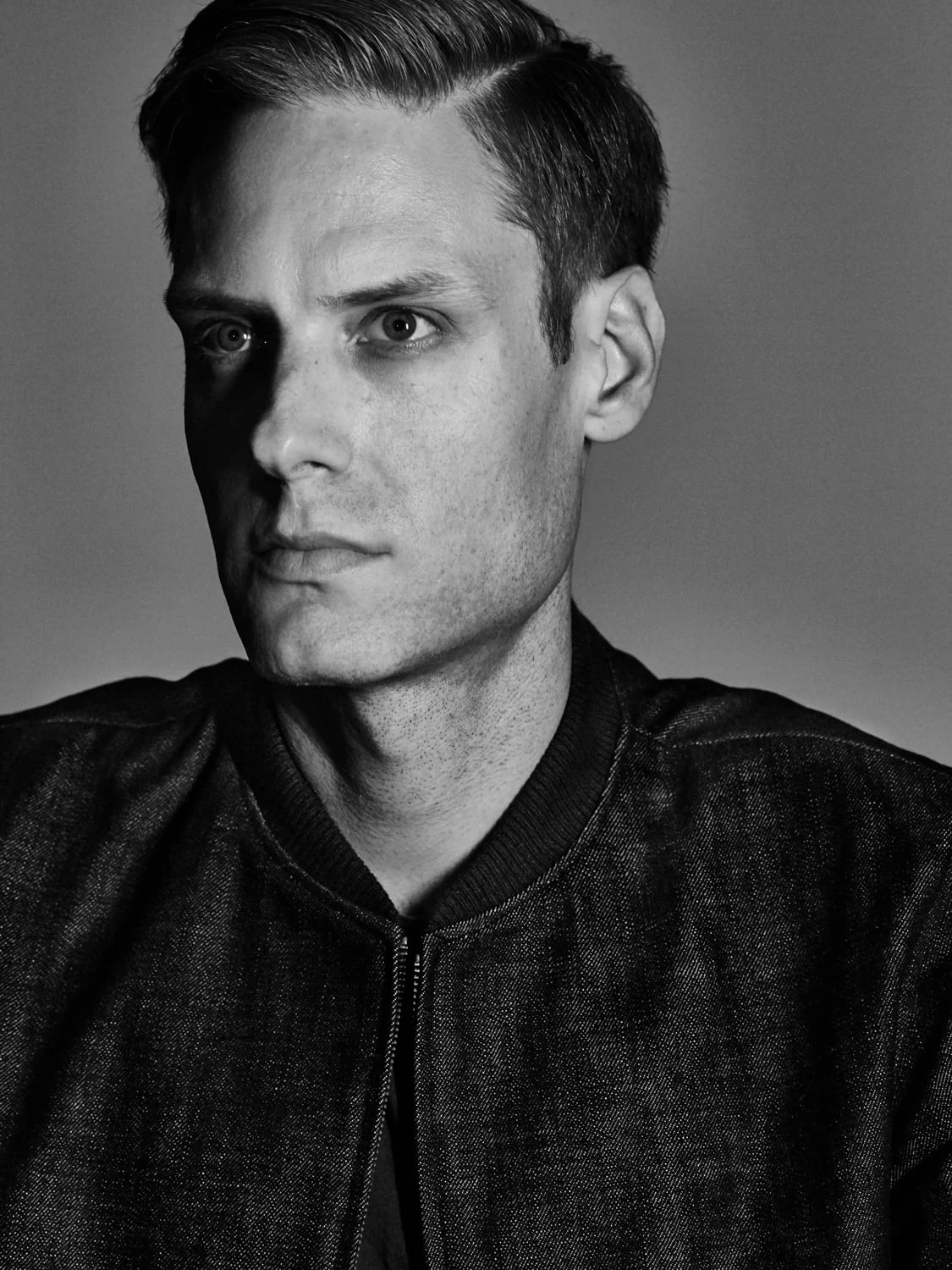 Black and white portrait of a man with slicked side-parted hair wearing a dark denim bomber jacket against a grey backdrop.
