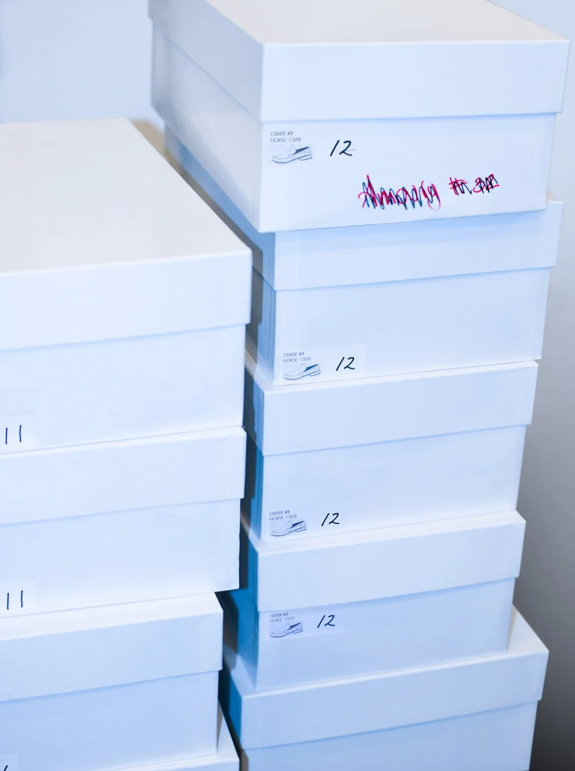 Stacks of white shoe boxes with size labels and shoe sketches, one marked in pink marker, against a pale wall.