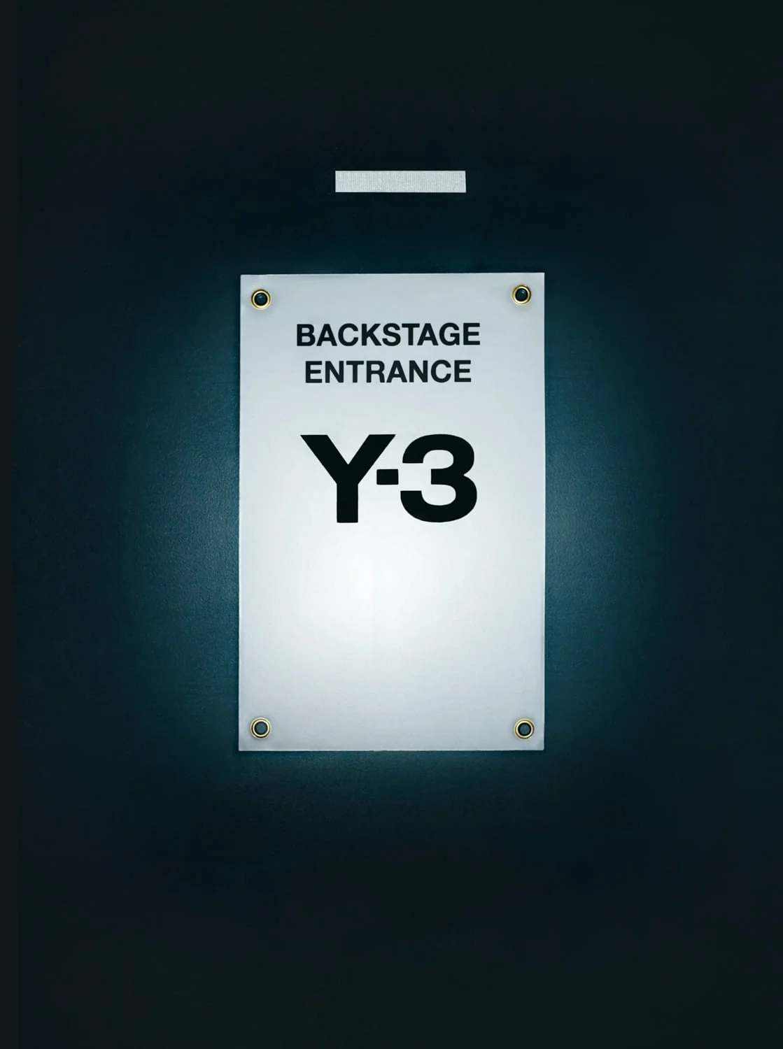 White Backstage Entrance sign with the Y-3 logo, fixed by brass grommets to a dark wall under a spotlight.