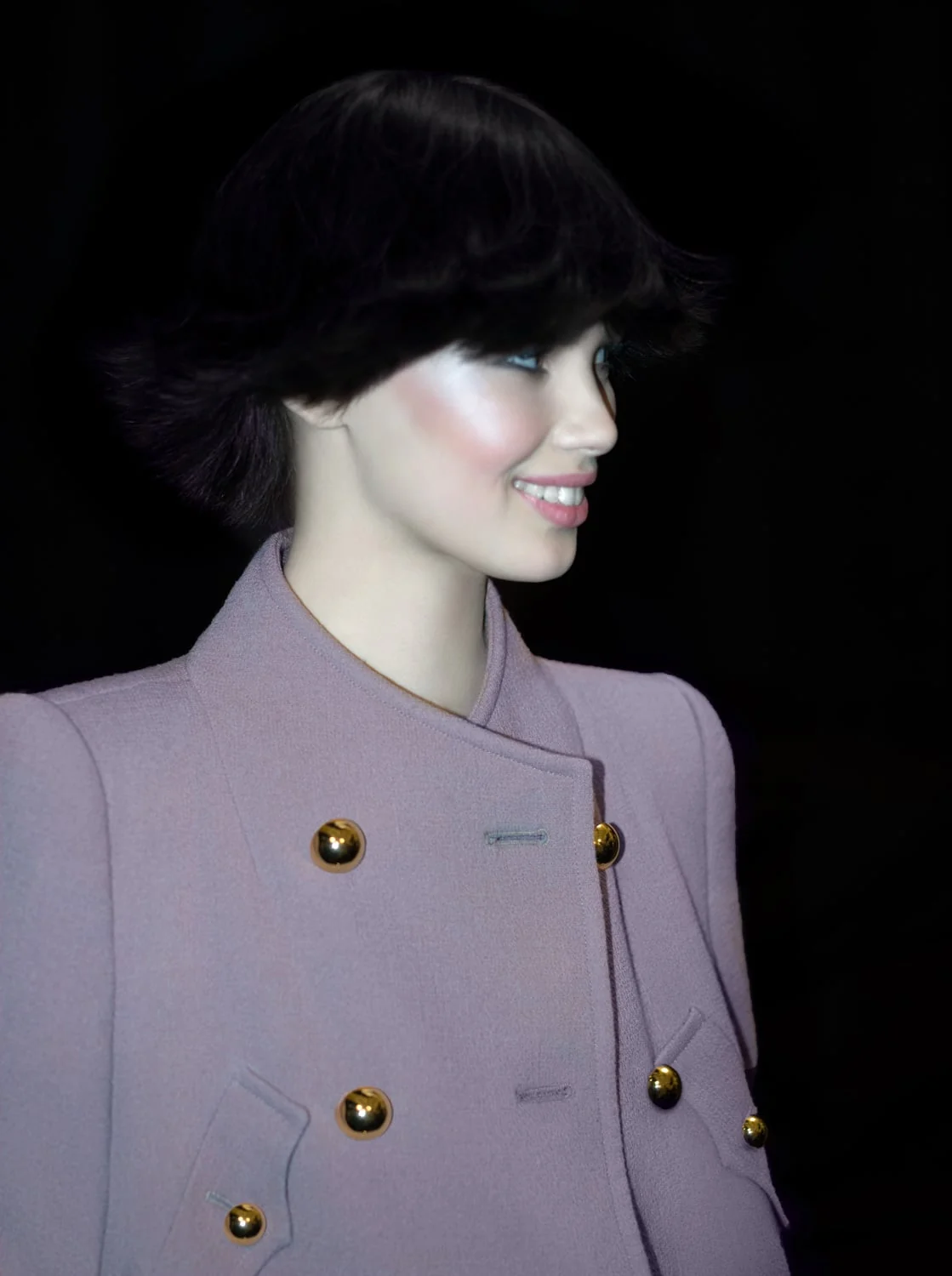 Smiling model with a dark bob in a lavender double-breasted coat with gold buttons, against a black background.