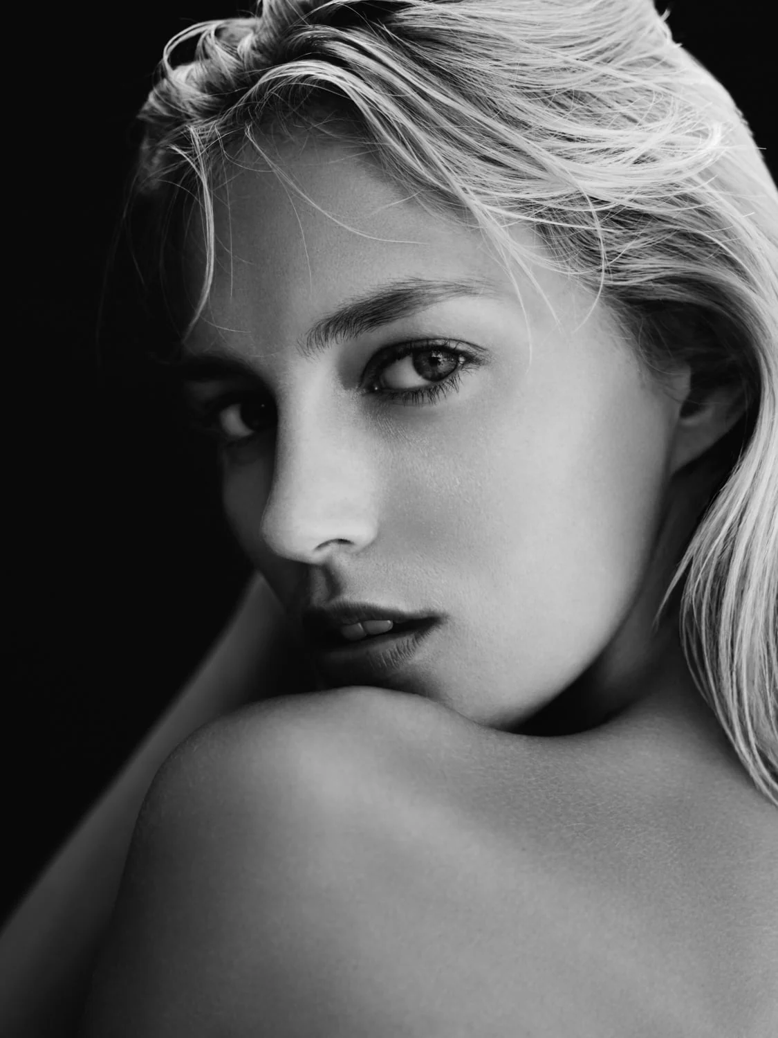 Black and white close-up of a blonde woman looking back over her bare shoulder against a dark background.