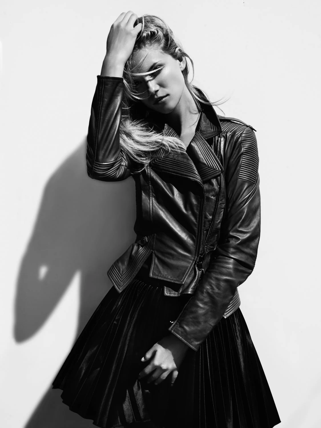 Black and white portrait of Sonya Gorelova in a leather biker jacket and pleated skirt, hand in her hair against a white wall.