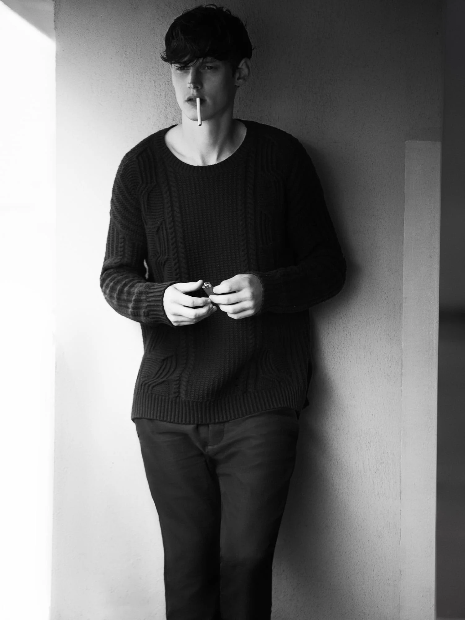 Black-and-white portrait of Adam Butcher in a dark cable-knit sweater, cigarette in mouth, leaning against a wall.