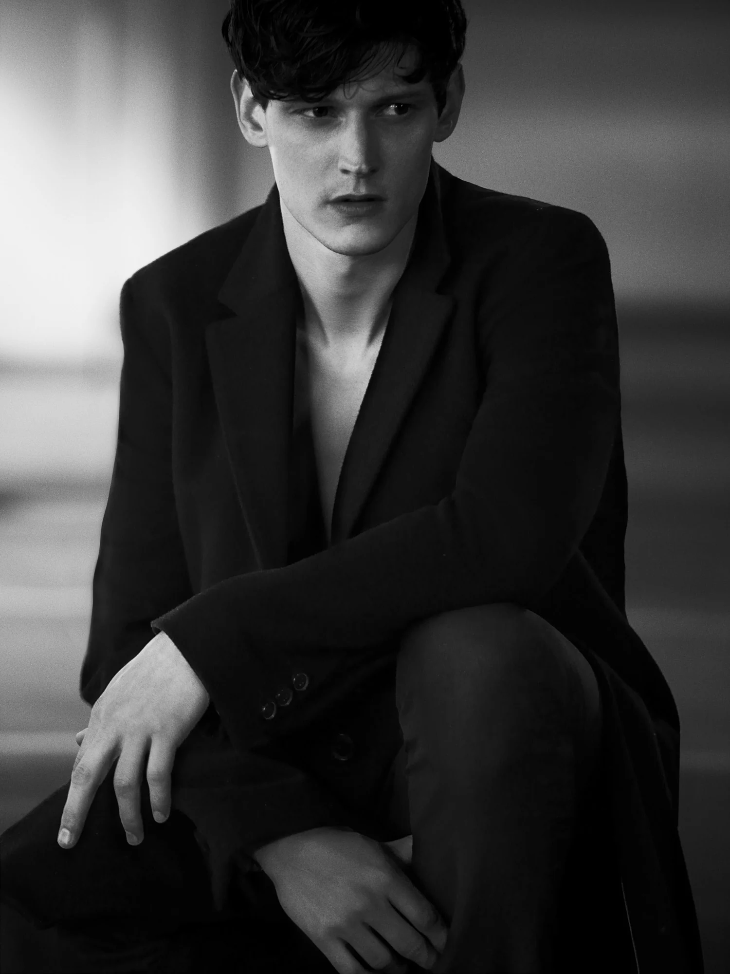 Black and white portrait of Adam Butcher seated in a dark open coat, bare chest, glancing to the side.