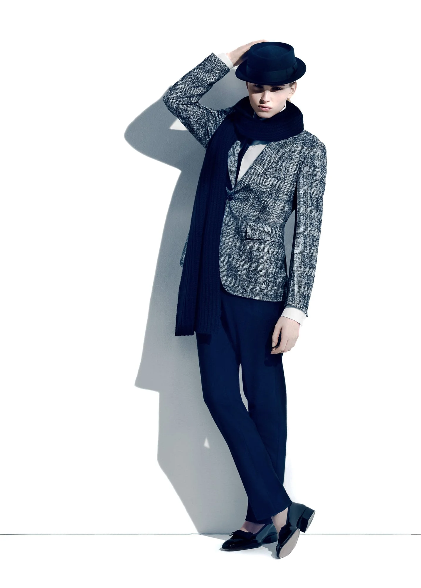 Model in a grey tweed blazer, navy scarf, trousers and fedora leaning against a white wall, for Apropos.