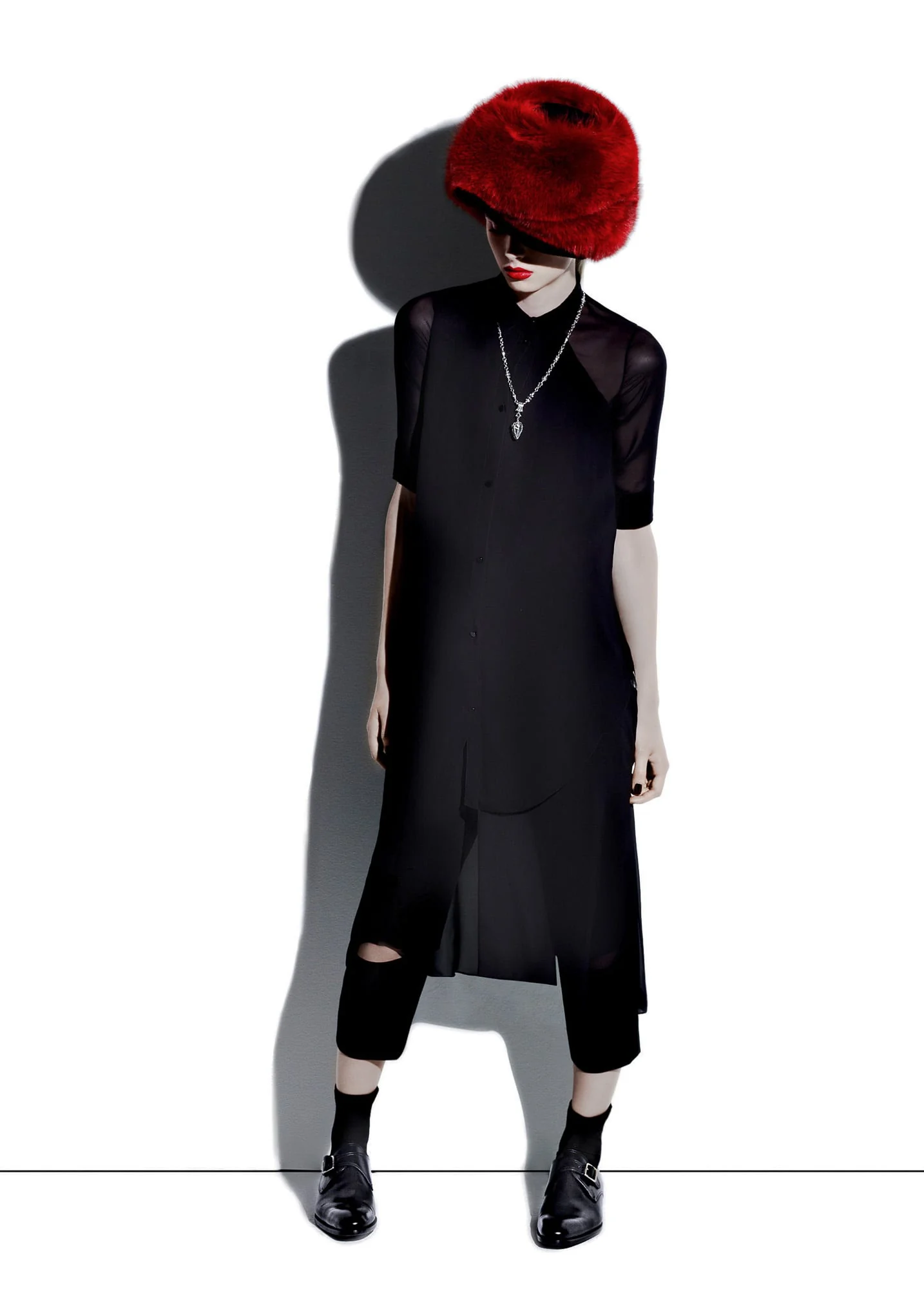 Model in a red fur hat, sheer black tunic, cropped trousers and buckled shoes against a white wall, for Apropos.