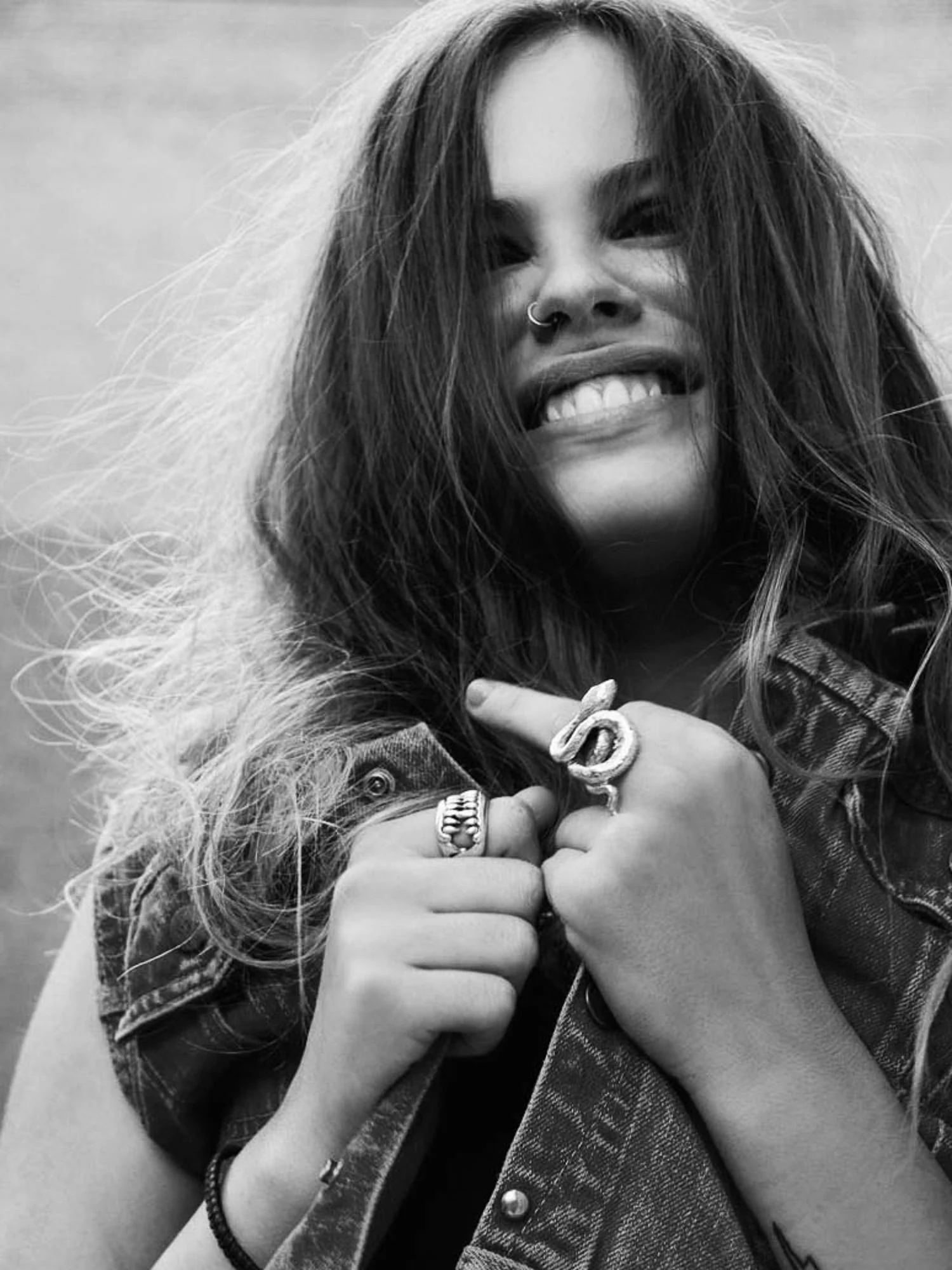 Black-and-white portrait of Atlanta de Cadenet Taylor laughing, hair windswept, gripping a denim vest, wearing silver rings.