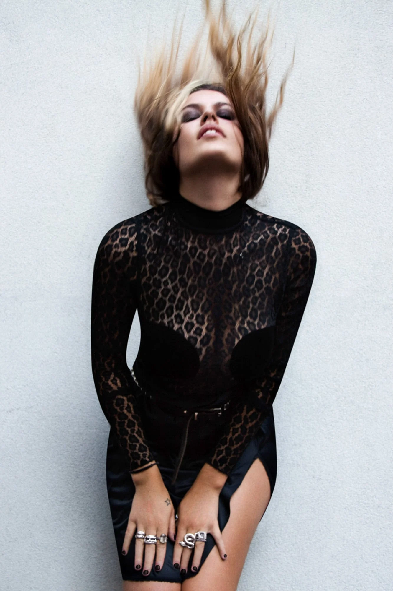 Atlanta de Cadenet Taylor tossing her hair in a sheer black leopard lace top against a white wall.