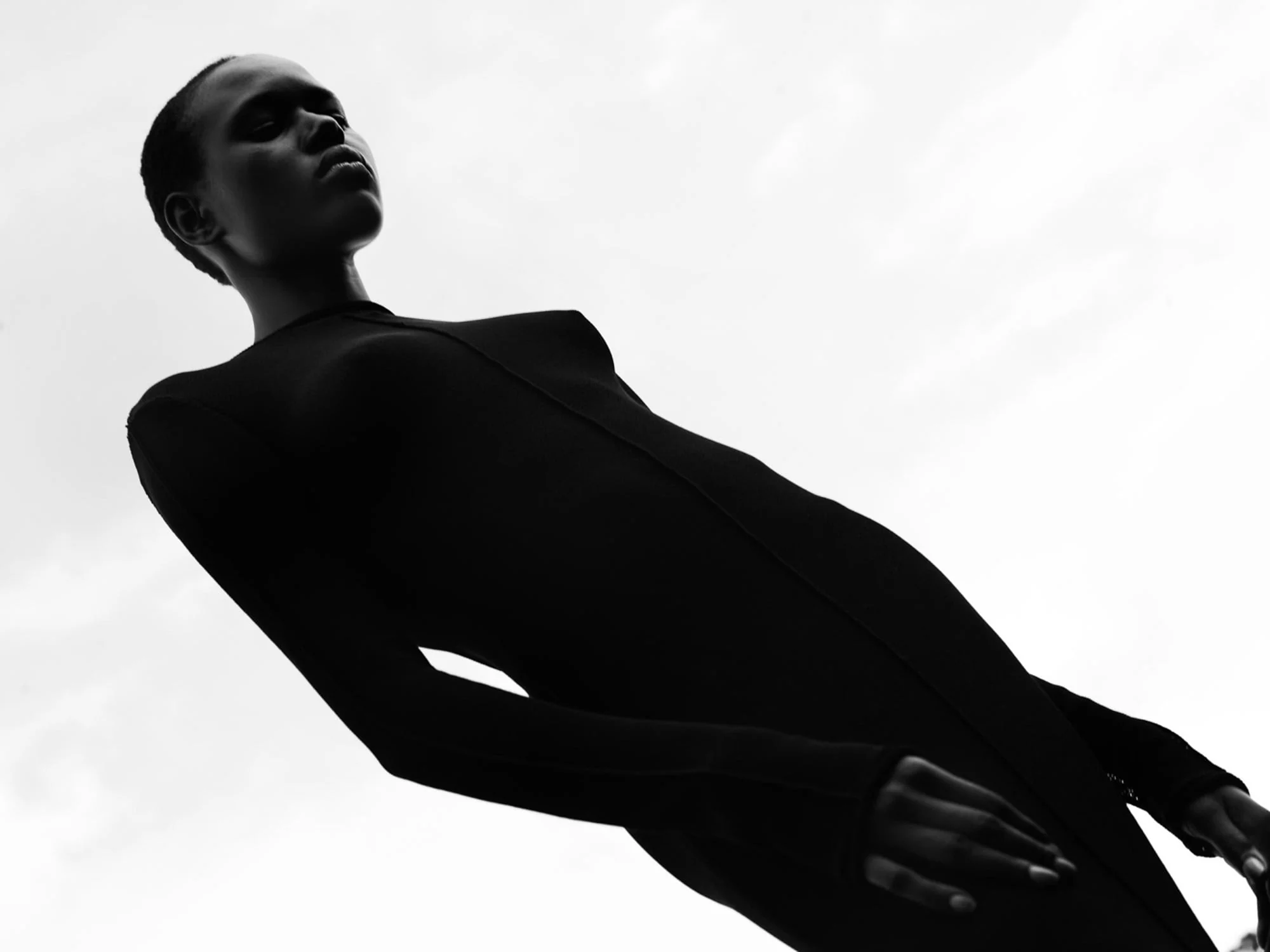 Model in a fitted black long-sleeve dress shot from below against a pale sky, in black and white, for Bon.